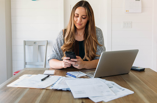 A woman sitting at a table with her phone, laptop and papers.